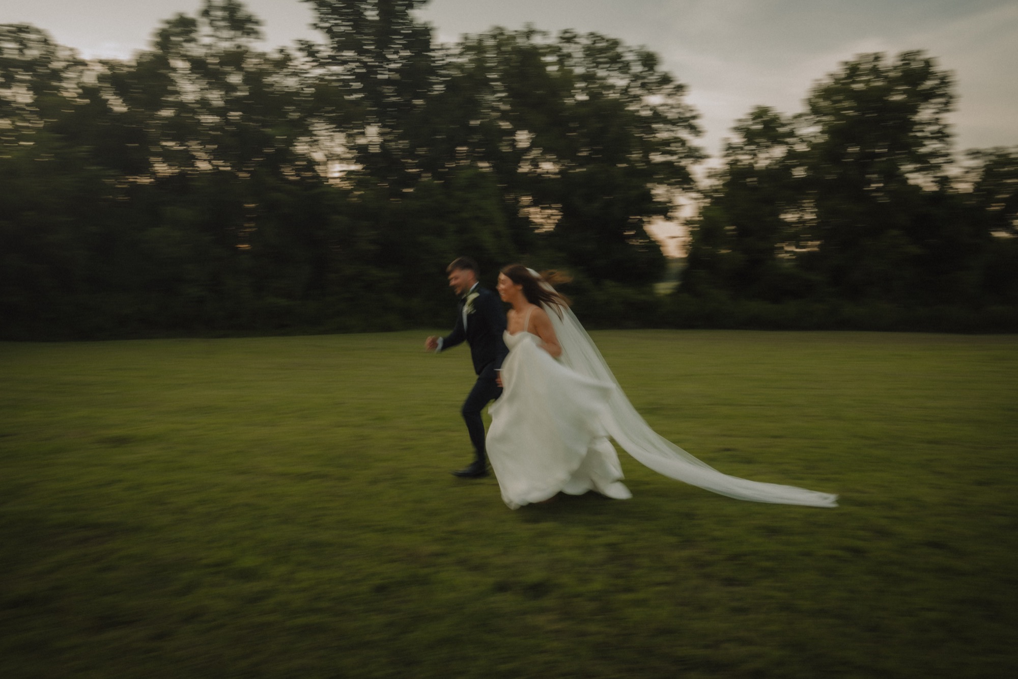 Bride and groom running hand in hand across an open field at dusk, veil streaming behind them in motion blur